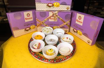 assorted bengali sweets in white bowls displayed on a decorative tray with gift boxes in the background